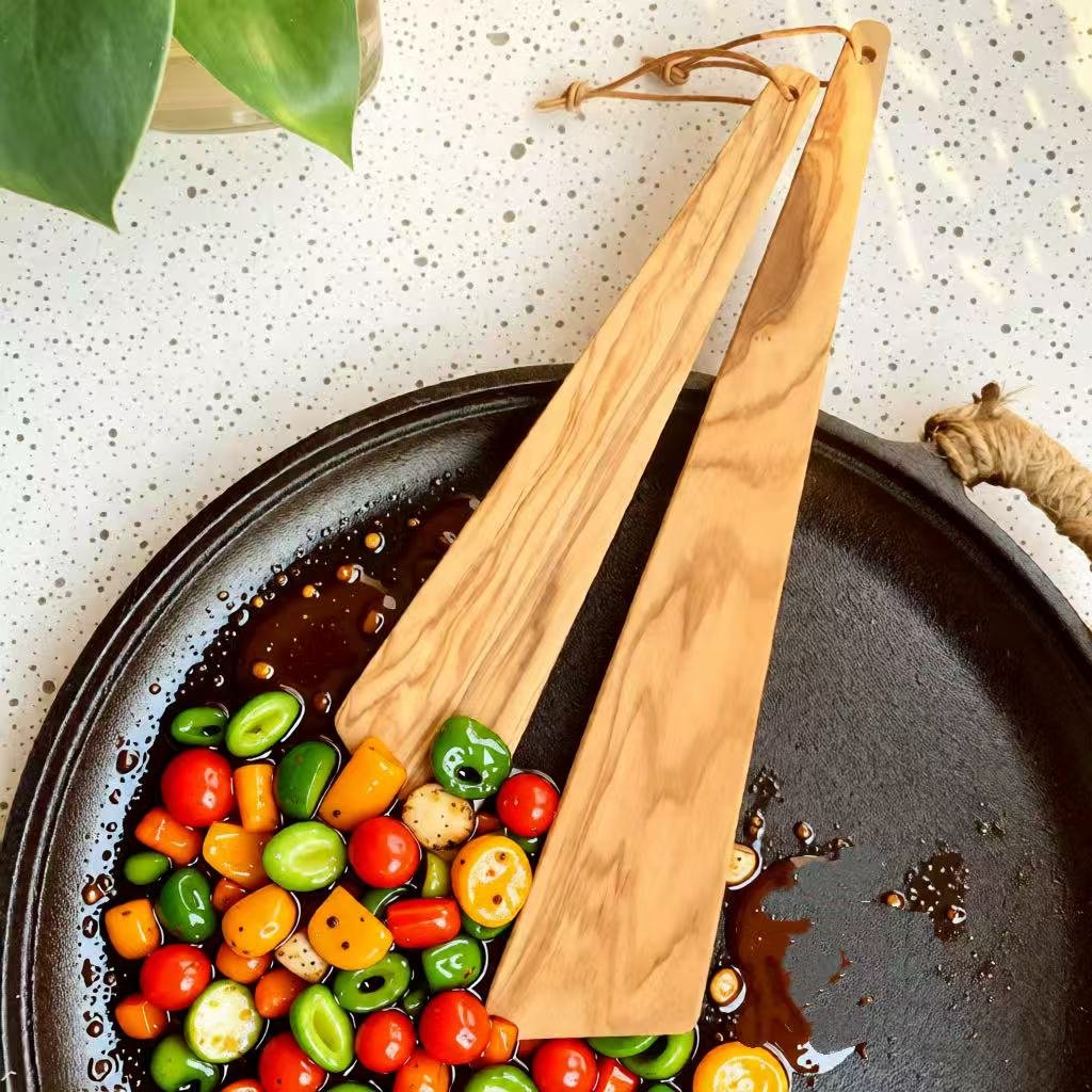 Close-up of olive wood triangle spatula with thin edge resting on black pan with colorful vegetables, highlighting natural wood grain and portable design