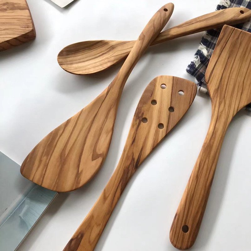 A set of handcrafted olive wood kitchen utensils including a solid spoon, slotted spatula, and flat turner, arranged on a white surface with a checkered cloth accent
